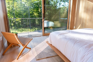 A modern bedroom features a large glass door opening to a balcony with a view of lush green trees. The room has a minimalistic design with wooden elements, including a woven chair and light wooden flooring. A white bedspread covers the neatly made bed, and sunlight filters through the glass, casting soft shadows on the floor.