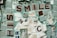 A mosaic artwork composed of variously colored tiles in beige, green, and blue shades. The tiles are arranged to form partial words such as 'SMILE' and 'HIS'. Textured patterns and chipped surfaces add a rustic feel. There is a small image of a person on one of the tiles.