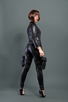 A person wearing a sleek, black textured outfit with high heels and a stylish bob haircut. They are holding a weapon and have tactical gear strapped to their legs, conveying a sense of readiness and confidence.