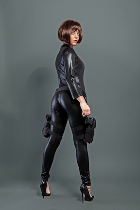 A person wearing a sleek, black textured outfit with high heels and a stylish bob haircut. They are holding a weapon and have tactical gear strapped to their legs, conveying a sense of readiness and confidence.
