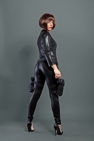 A person wearing a sleek, black textured outfit with high heels and a stylish bob haircut. They are holding a weapon and have tactical gear strapped to their legs, conveying a sense of readiness and confidence.