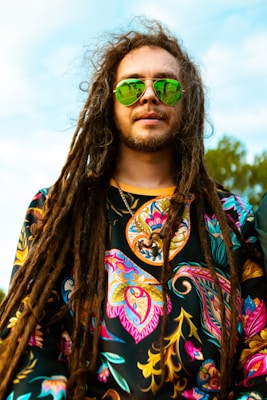 A person with long dreadlocks and a beard wears reflective green sunglasses and a colorful, patterned shirt. The background shows a clear sky with some greenery.