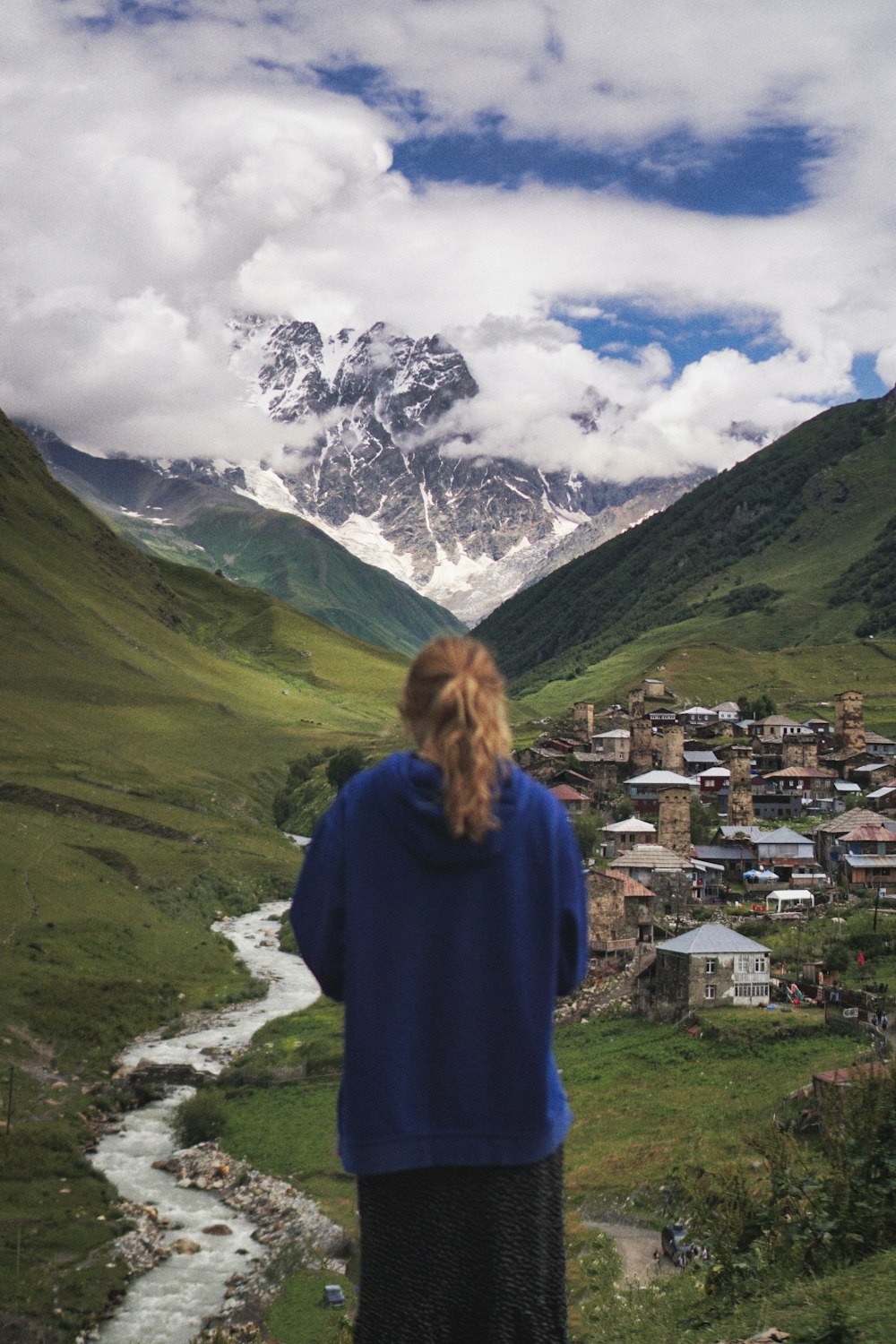 woman in blue jacket standing on green grass field near village during daytime