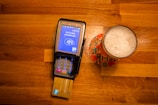 A payment terminal with a card inserted is placed on a wooden table next to a glass of beer on a colorful coaster. The terminal screen displays a payment request for 1.91 EUR.