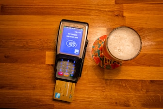 A payment terminal with a card inserted is placed on a wooden table next to a glass of beer on a colorful coaster. The terminal screen displays a payment request for 1.91 EUR.