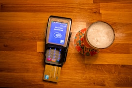 A payment terminal with a card inserted is placed on a wooden table next to a glass of beer on a colorful coaster. The terminal screen displays a payment request for 1.91 EUR.