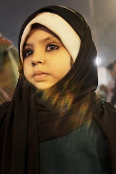 A young child wearing a black headscarf with a white under-scarf gazes thoughtfully into the distance. Light reflections create a lens flare effect across the image, giving it a warm tone.