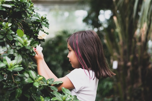 A young girl with dark hair and a pink streak is gently reaching towards a white flower amidst a lush green garden. She appears focused and curious, surrounded by dense foliage and natural serene light.