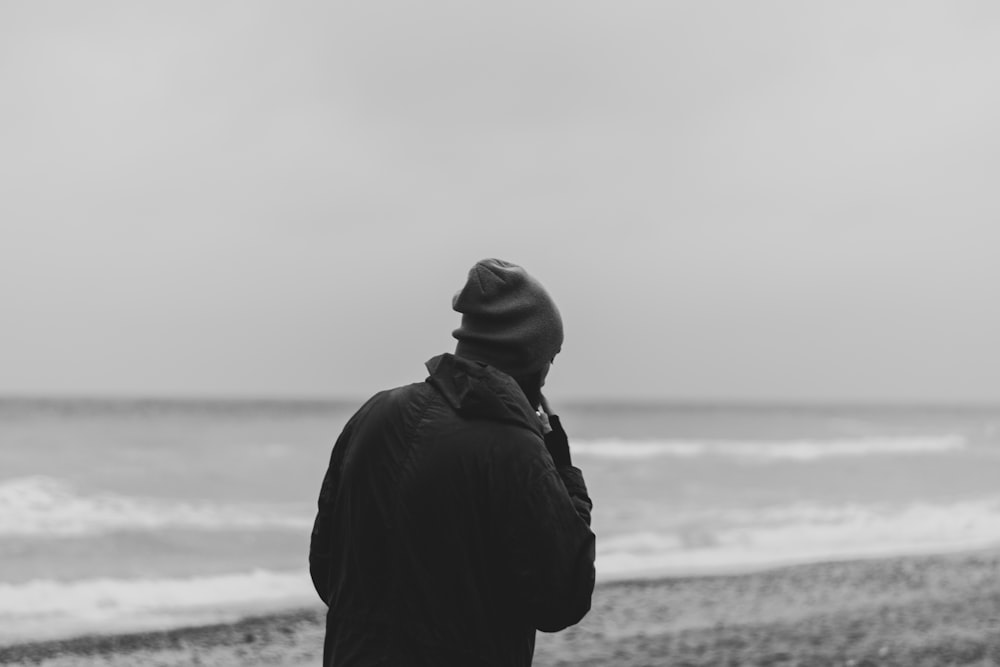grayscale photo of person in hoodie standing on seashore
