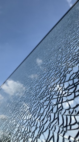 A cracked glass surface against a backdrop of a clear blue sky with a few clouds. The glass shows intricate patterns and reflections, giving an abstract aesthetic.