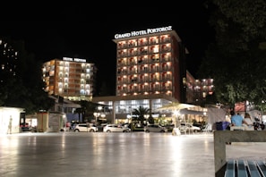 A well-lit hotel complex at night with prominent signage for Grand Hotel Portorož and Hotel Neptun. The buildings are illuminated with warm lights, and cars are parked in front of the hotel. Several people are visible, some seated and others walking by, under a clear night sky.
