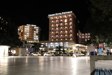 A well-lit hotel complex at night with prominent signage for Grand Hotel Portorož and Hotel Neptun. The buildings are illuminated with warm lights, and cars are parked in front of the hotel. Several people are visible, some seated and others walking by, under a clear night sky.