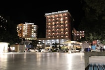 A well-lit hotel complex at night with prominent signage for Grand Hotel Portorož and Hotel Neptun. The buildings are illuminated with warm lights, and cars are parked in front of the hotel. Several people are visible, some seated and others walking by, under a clear night sky.