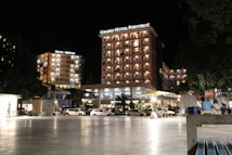 A well-lit hotel complex at night with prominent signage for Grand Hotel Portorož and Hotel Neptun. The buildings are illuminated with warm lights, and cars are parked in front of the hotel. Several people are visible, some seated and others walking by, under a clear night sky.