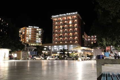 A well-lit hotel complex at night with prominent signage for Grand Hotel Portorož and Hotel Neptun. The buildings are illuminated with warm lights, and cars are parked in front of the hotel. Several people are visible, some seated and others walking by, under a clear night sky.