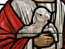 A detailed depiction of a stained glass artwork featuring a lamb held by a pair of hands. The scene is framed with bold black outlines against textured backgrounds in red, white, and beige hues.