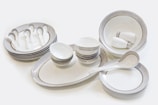 A set of neatly arranged ceramic tableware, consisting of plates, bowls, saucers, and spoons. The dishes are predominantly white with subtle gold and silver decorative bands around their edges. The bowls are stacked in varying sizes and the plates are similarly organized. Two small cups are placed inside a large bowl.