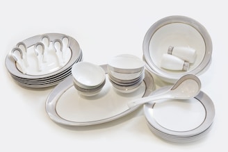 A set of neatly arranged ceramic tableware, consisting of plates, bowls, saucers, and spoons. The dishes are predominantly white with subtle gold and silver decorative bands around their edges. The bowls are stacked in varying sizes and the plates are similarly organized. Two small cups are placed inside a large bowl.