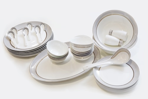 A set of neatly arranged ceramic tableware, consisting of plates, bowls, saucers, and spoons. The dishes are predominantly white with subtle gold and silver decorative bands around their edges. The bowls are stacked in varying sizes and the plates are similarly organized. Two small cups are placed inside a large bowl.