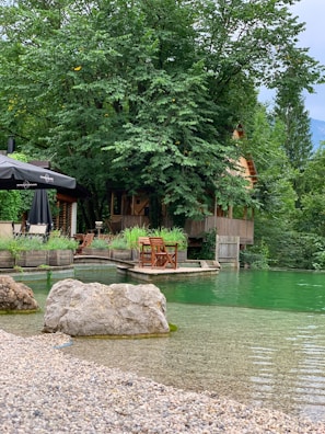 A tranquil lakeside scene featuring a rustic wooden cabin nestled among lush green trees. The cabin sits on a wooden deck with outdoor furniture, including chairs and tables. A small rocky shore with clear, calm water in shades of green is visible in the foreground. An umbrella provides shade over part of the deck area.