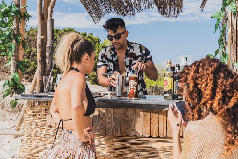 A beachside bar scene with a bartender preparing drinks for two women. The bartender is wearing sunglasses and a patterned shirt, and various bottles of alcohol are visible on the counter. The women are dressed in summer attire, one with a phone, and the setting includes tropical decorations and scenery.