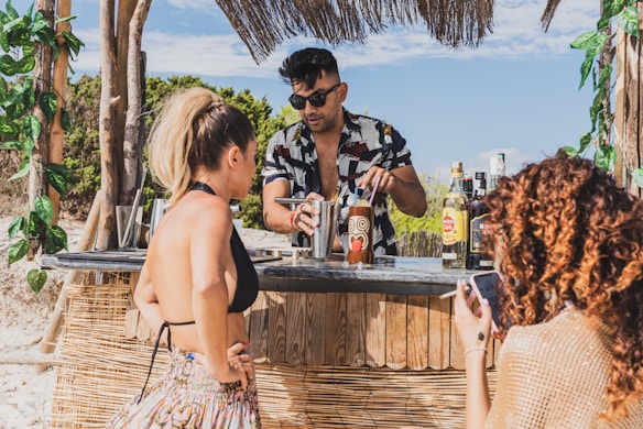 A beachside bar scene with a bartender preparing drinks for two women. The bartender is wearing sunglasses and a patterned shirt, and various bottles of alcohol are visible on the counter. The women are dressed in summer attire, one with a phone, and the setting includes tropical decorations and scenery.