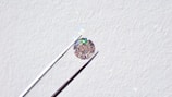 A single round diamond is delicately held by a pair of metal tweezers against a textured white background. The diamond sparkles with a spectrum of colors, highlighting its cut and clarity.