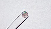 A single round diamond is delicately held by a pair of metal tweezers against a textured white background. The diamond sparkles with a spectrum of colors, highlighting its cut and clarity.