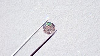 A single round diamond is delicately held by a pair of metal tweezers against a textured white background. The diamond sparkles with a spectrum of colors, highlighting its cut and clarity.
