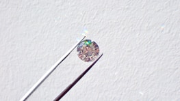 A single round diamond is delicately held by a pair of metal tweezers against a textured white background. The diamond sparkles with a spectrum of colors, highlighting its cut and clarity.