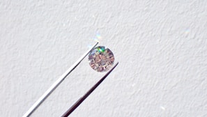 A single round diamond is delicately held by a pair of metal tweezers against a textured white background. The diamond sparkles with a spectrum of colors, highlighting its cut and clarity.