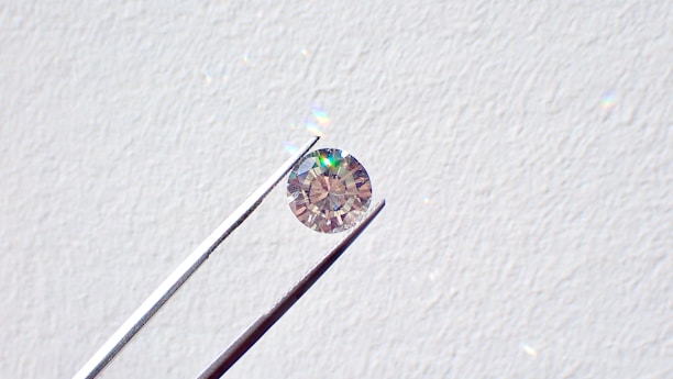 A single round diamond is delicately held by a pair of metal tweezers against a textured white background. The diamond sparkles with a spectrum of colors, highlighting its cut and clarity.
