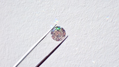 A single round diamond is delicately held by a pair of metal tweezers against a textured white background. The diamond sparkles with a spectrum of colors, highlighting its cut and clarity.