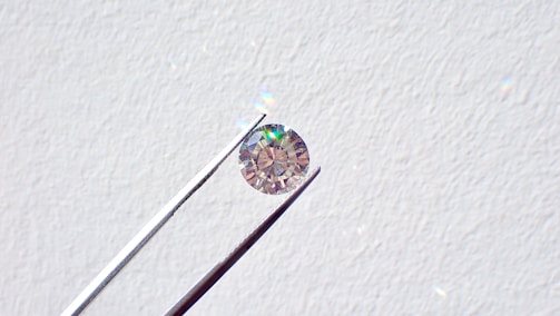 A single round diamond is delicately held by a pair of metal tweezers against a textured white background. The diamond sparkles with a spectrum of colors, highlighting its cut and clarity.