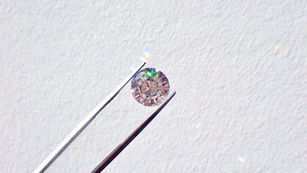 A single round diamond is delicately held by a pair of metal tweezers against a textured white background. The diamond sparkles with a spectrum of colors, highlighting its cut and clarity.