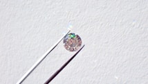 A single round diamond is delicately held by a pair of metal tweezers against a textured white background. The diamond sparkles with a spectrum of colors, highlighting its cut and clarity.