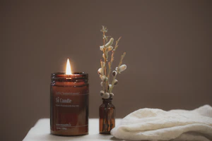 A lit candle in an amber jar beside dried flowers on a linen-draped table