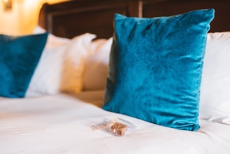 A neatly made bed with a set of blue velvet throw pillows and a small packaged brownie placed on the white bedspread. The background features a wooden headboard and soft, warm lighting, suggesting a cozy and inviting atmosphere.