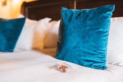 A neatly made bed with a set of blue velvet throw pillows and a small packaged brownie placed on the white bedspread. The background features a wooden headboard and soft, warm lighting, suggesting a cozy and inviting atmosphere.