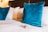 A neatly made bed with a set of blue velvet throw pillows and a small packaged brownie placed on the white bedspread. The background features a wooden headboard and soft, warm lighting, suggesting a cozy and inviting atmosphere.