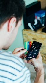 A person is holding a smartphone in one hand displaying a banking or finance application and a colorful credit card in the other hand. Nearby, a shelf holds various gaming consoles and accessories.