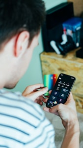 A person is holding a smartphone in one hand displaying a banking or finance application and a colorful credit card in the other hand. Nearby, a shelf holds various gaming consoles and accessories.