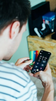 A person is holding a smartphone in one hand displaying a banking or finance application and a colorful credit card in the other hand. Nearby, a shelf holds various gaming consoles and accessories.