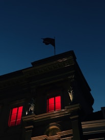 The image depicts a building with classical architectural elements, featuring columns and detailed molding. Two windows are illuminated with a striking red light. A flag is positioned atop the building against a dark blue evening sky.