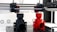 A 3D printer is in the process of printing two objects, one in black and one in red. The setup includes two extruder heads that are actively working on creating these cylindrical, industrial-like components. The printer is situated on a clean, white counter with visible branding and technical details in the background.