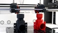 A 3D printer is in the process of printing two objects, one in black and one in red. The setup includes two extruder heads that are actively working on creating these cylindrical, industrial-like components. The printer is situated on a clean, white counter with visible branding and technical details in the background.