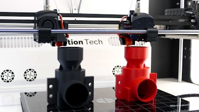 A 3D printer is in the process of printing two objects, one in black and one in red. The setup includes two extruder heads that are actively working on creating these cylindrical, industrial-like components. The printer is situated on a clean, white counter with visible branding and technical details in the background.