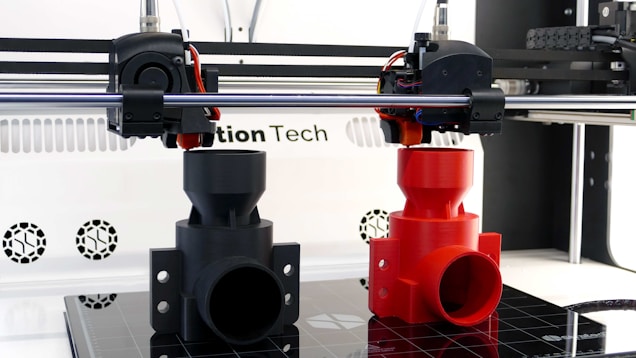A 3D printer is in the process of printing two objects, one in black and one in red. The setup includes two extruder heads that are actively working on creating these cylindrical, industrial-like components. The printer is situated on a clean, white counter with visible branding and technical details in the background.