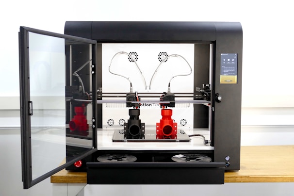 A modern 3D printer with an open door on the left, showcasing two spools of filament inside. The device is positioned on a wooden surface, with a black and metallic structure. The interior includes tubes and a printing head, with visible red and black filament on the printing platform.