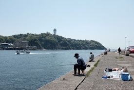 People are fishing along a concrete pier with a view of a body of water. A boat is moving across the water, and in the background, a densely forested hill with buildings and a tall tower can be seen. Various objects, including bags and a cooler, are placed on the pier.